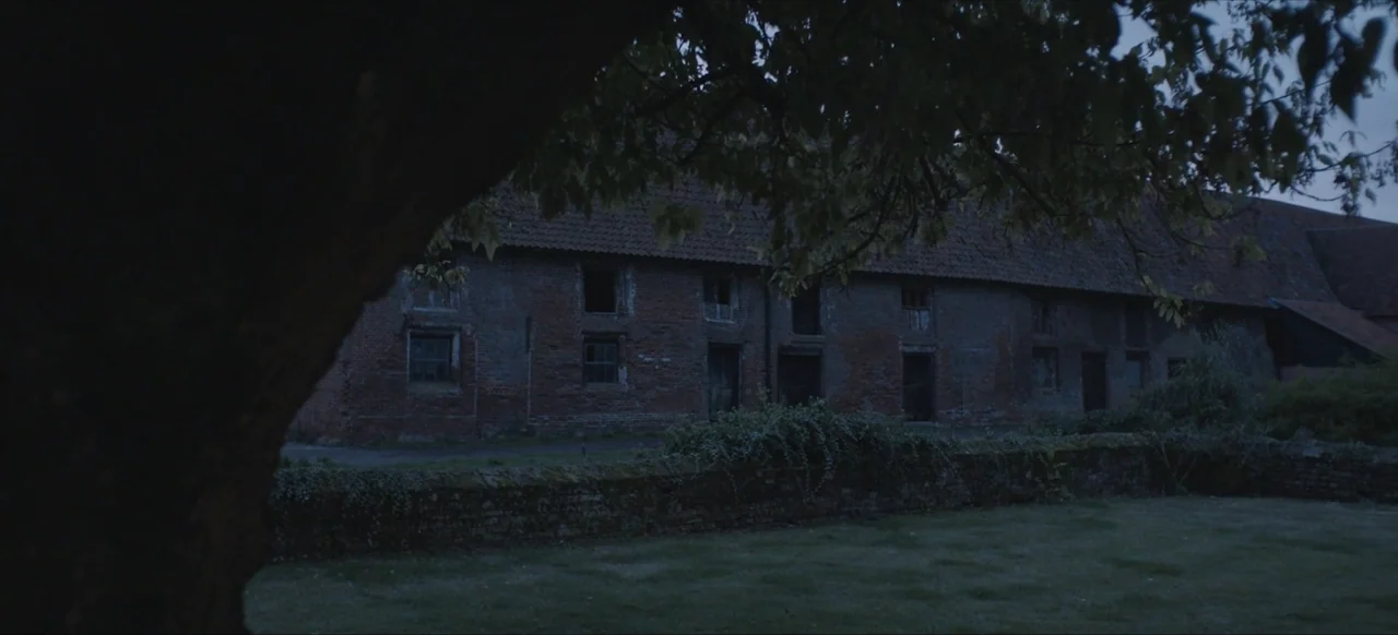 We Go In at Dawn (2020) movie still: establishing — An old red brick farm building with weathered windows sits behind a stone wall. The foreground…