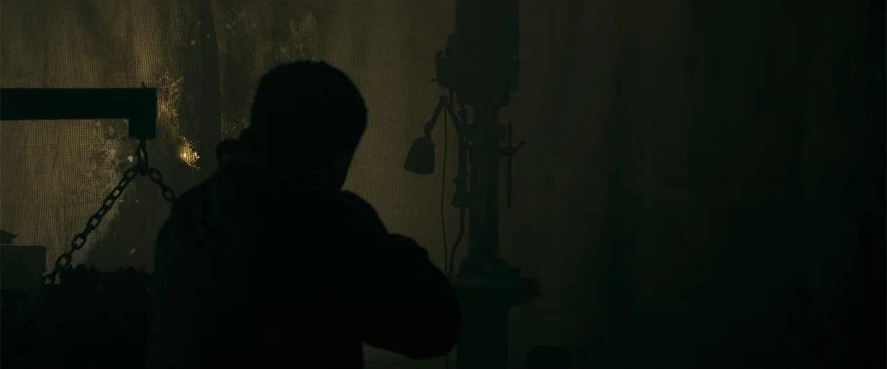 The Beekeeper (2024) movie still: medium — A dark workshop space features a drill press silhouetted against a textured mesh window. An individual…