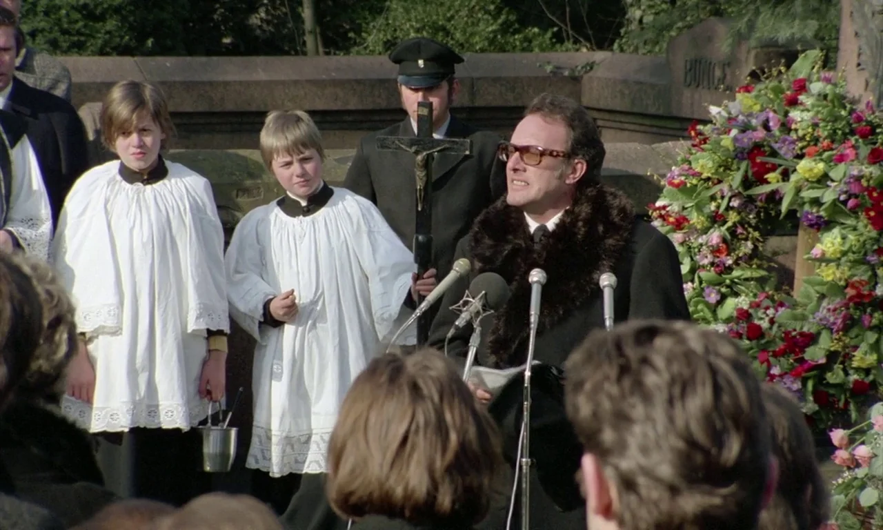 The Lost Honour of Katharina Blum (1975) movie still: medium — A funeral setting with stone monuments and a large floral wreath in the background. A…