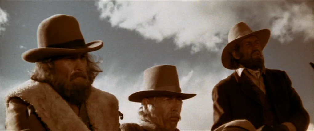 Pat Garrett & Billy The Kid (1973) movie still: medium — Three men wearing western hats stand against a bright blue sky with scattered clouds. The…