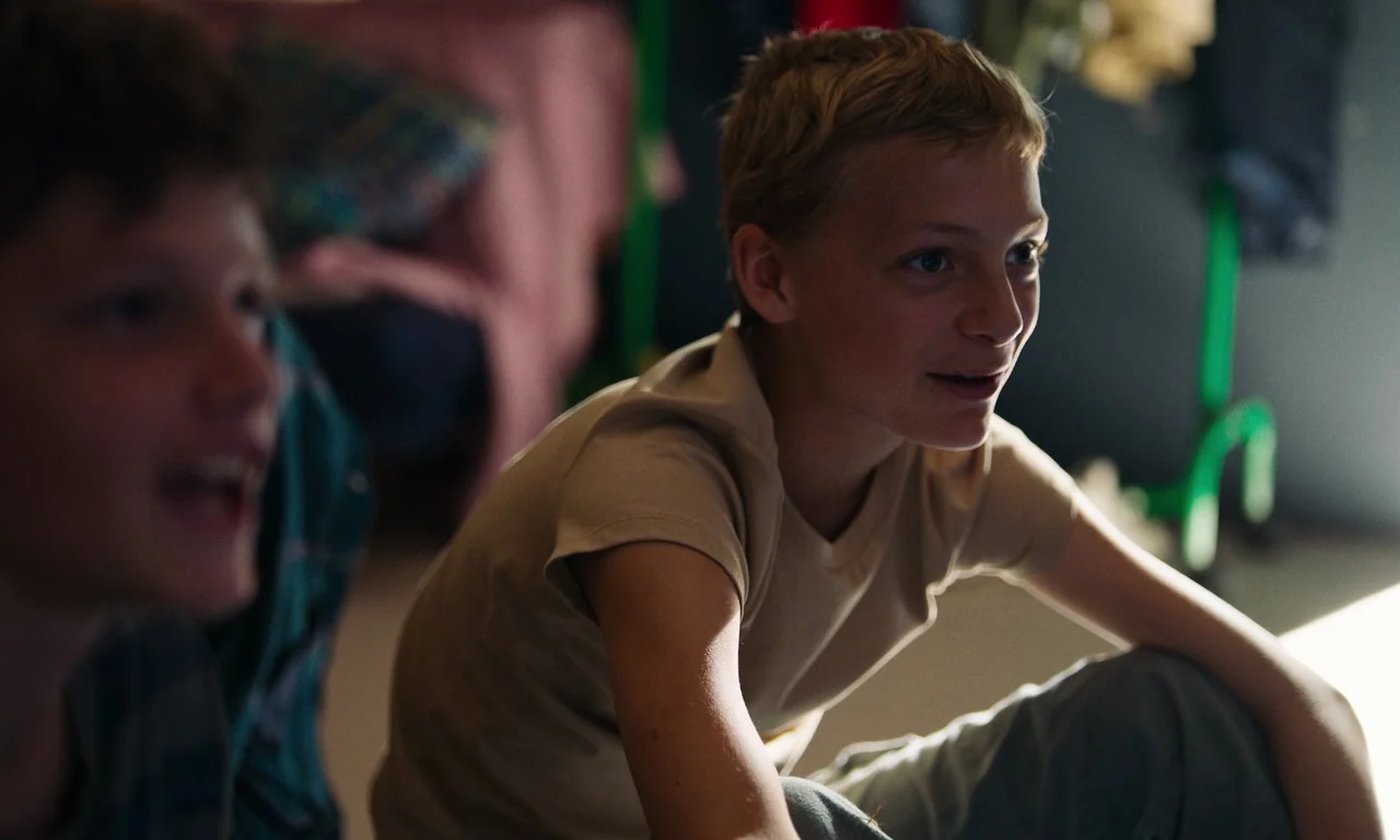 Close (2022) movie still: medium — A dimly lit room with a boy in a beige shirt sitting on the floor. A second boy is partially visible in the…