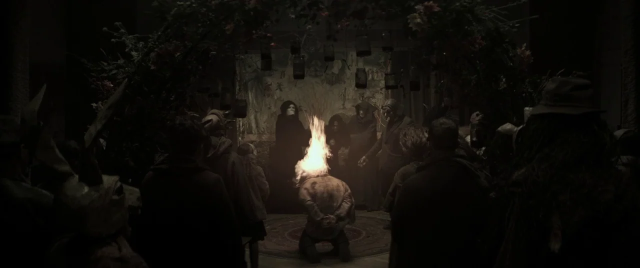 Lord of Misrule (2023) movie still: wide — A dimly lit room is decorated with hanging jars and an arched floral entrance. A group of masked figures…