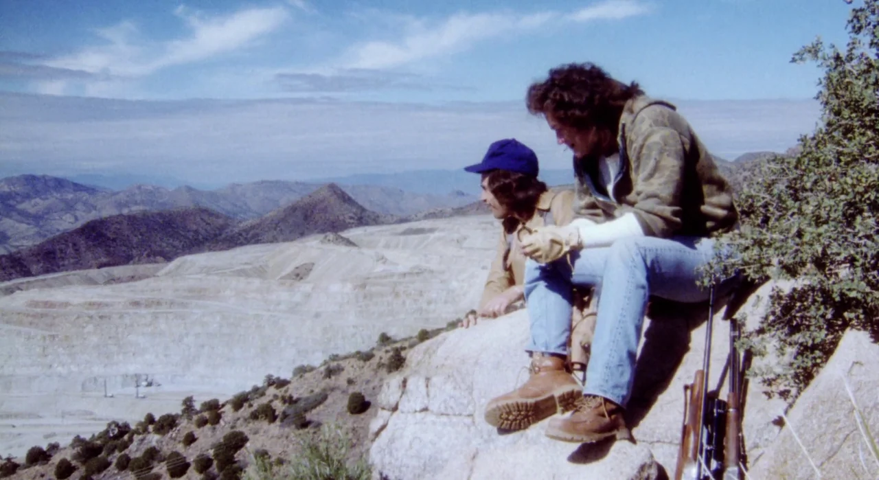 White Of The Eye (1987) movie still: fullbody — Two men sit atop a rocky cliff overlooking a vast open-pit mining site. The distant mountains…