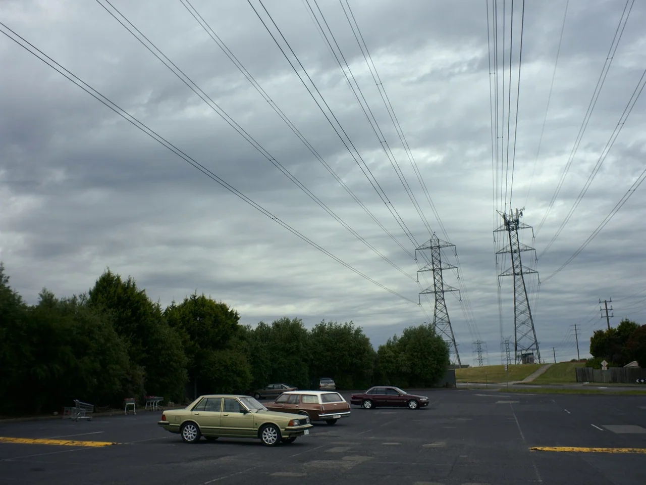 Of An Age (2022) movie still: wide — A quiet asphalt parking lot is dominated by three vintage vehicles and power pylons. Rows of overhead power…