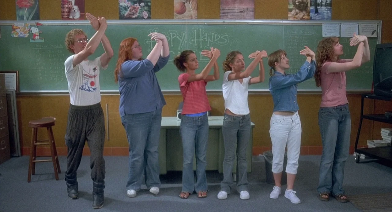 Napoleon Dynamite (2004) movie still: wide — A school room with a large green chalkboard covered in chalk writing and posters. Six people stand in a…