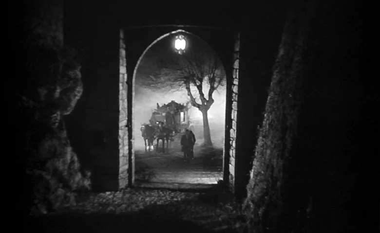 Black Sunday (1960) movie still: wide — A dark, arched stone gateway frames a fog-filled road at night. A horse-drawn carriage and a solitary figure…