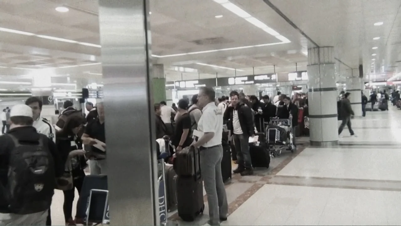 Vai Corinthians (2023) movie still: medium — A crowded airport terminal features polished floors and structural white columns. A man stands in the…