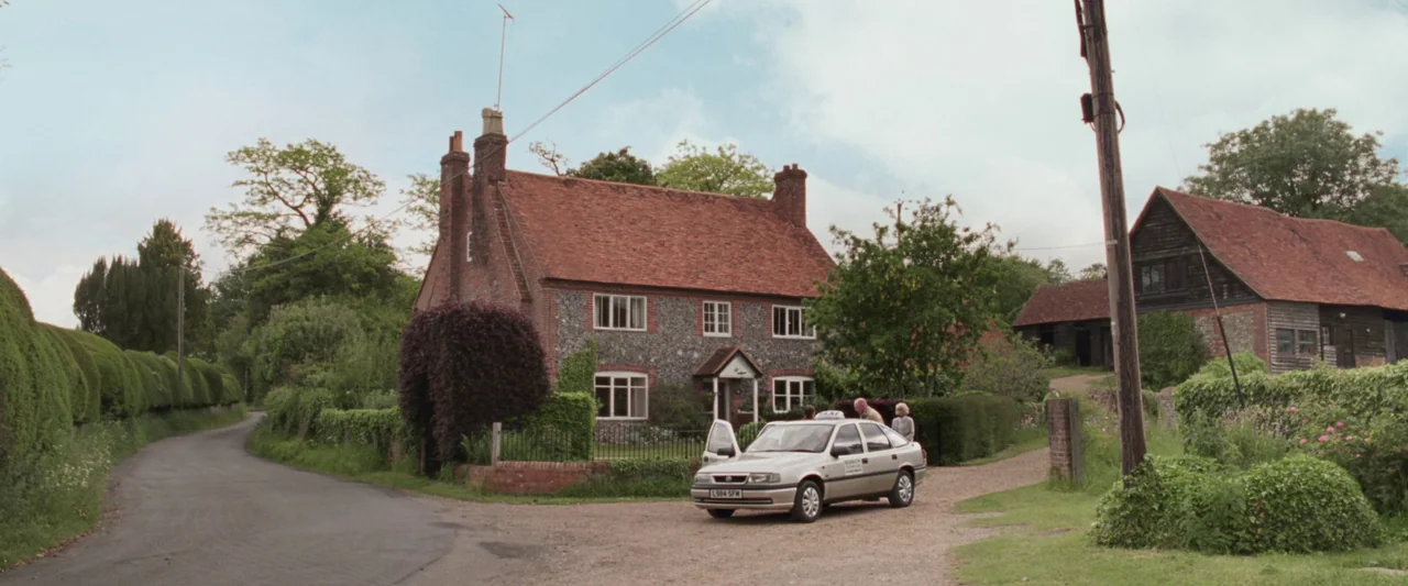 Last Night in Soho (2021) movie still: wide — A rural flint cottage with a tiled roof stands beside a quiet country road. A silver sedan taxi is…
