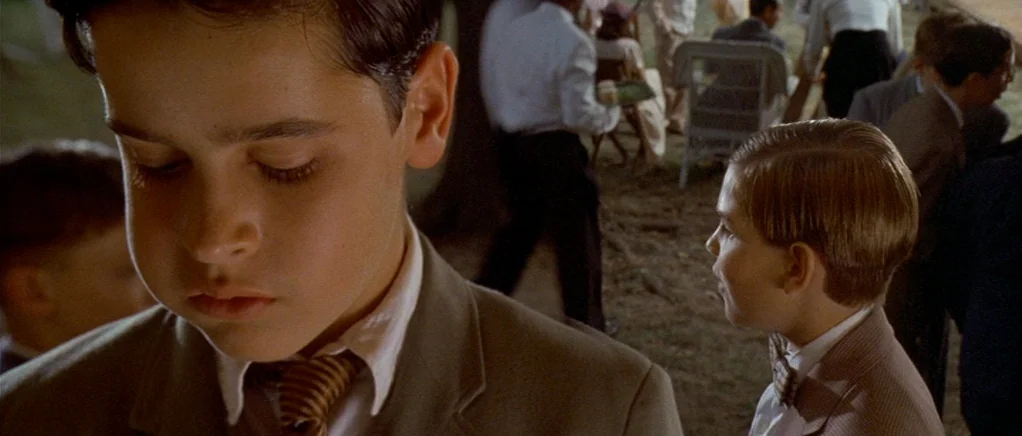 King of the Hill (1993) movie still: closeup — A group of children in 1940s formal wear stand on a grassy lawn. One boy in the foreground keeps his…