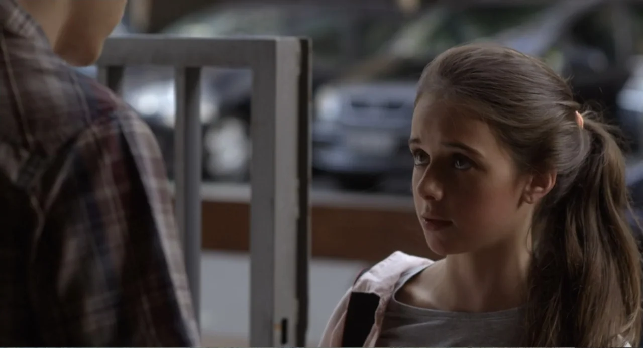Newton's Third Law (2017) movie still: medium — A sidewalk with a metal gate frame visible in the out-of-focus background. A teenage girl stands…