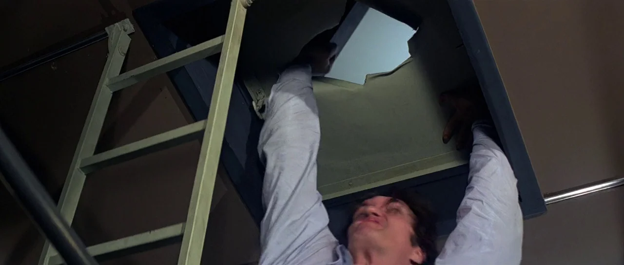 Moonraker (1979) movie still: medium — A man is underneath an open ceiling hatch leading to an upper level. He reaches upward with both hands to…