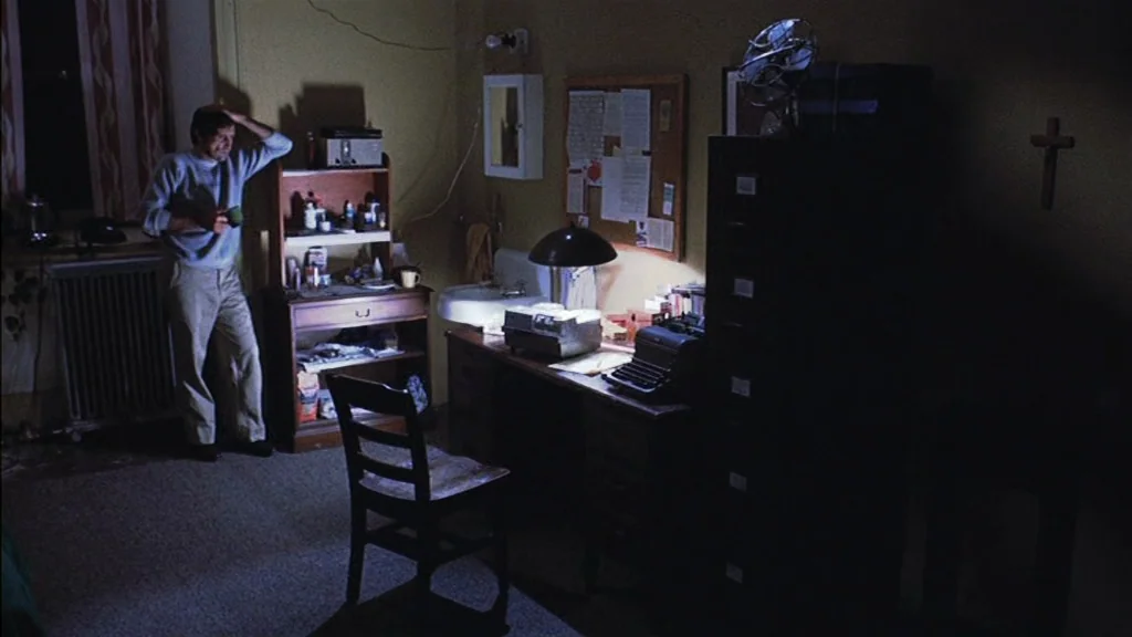 The Exorcist (1973) movie still: fullbody — A dimly lit room with plain walls, a desk with a lamp, and shelving units. A man stands in the shadow…