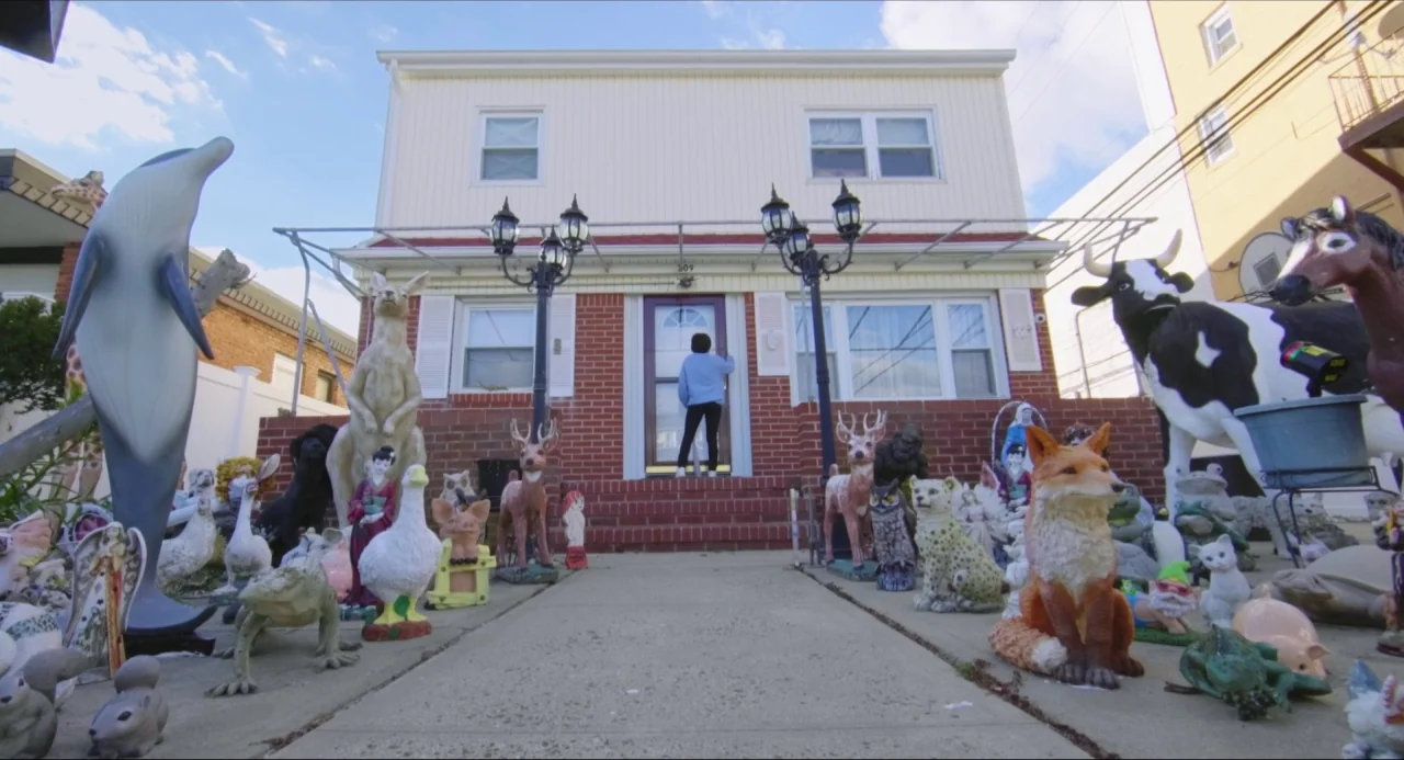 Like Me movie still: wide — A suburban house has a brick front porch crowded with many life-sized animal statues. A woman stands on the stairs…