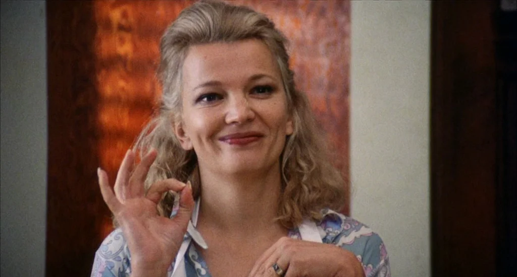 A Woman Under the Influence (1974) movie still: closeup — A woman stands in a home environment with wooden wall paneling behind her. She smiles…