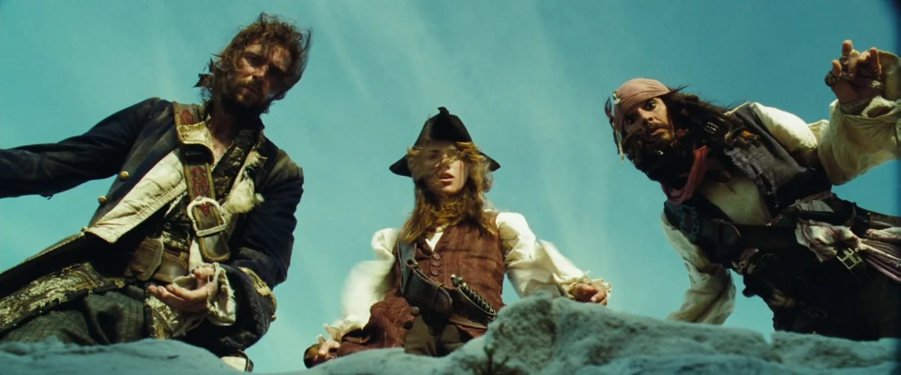 Pirates of the Caribbean: Dead Man’s Chest movie still: medium — A group of three pirates stand on a sandy beach under a clear blue sky. They look…