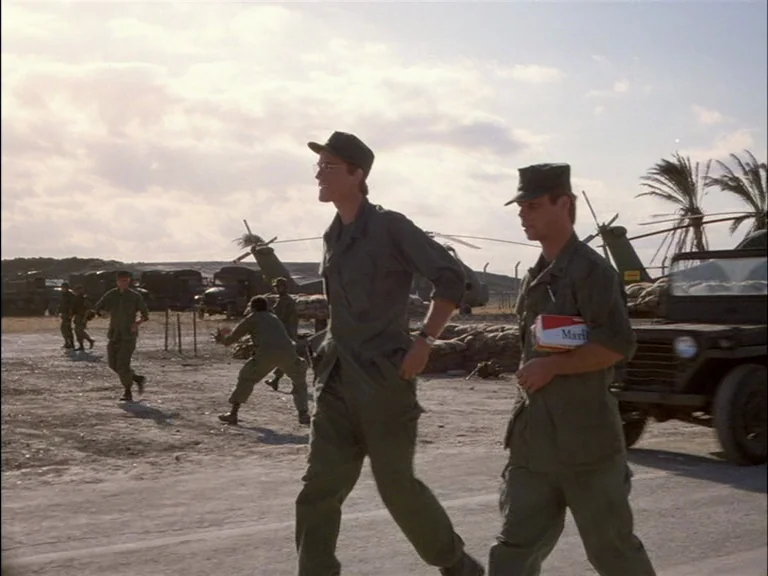 Full Metal Jacket (1987) movie still: fullbody — An arid military base is occupied by several soldiers, parked trucks, and helicopters. Two soldiers…