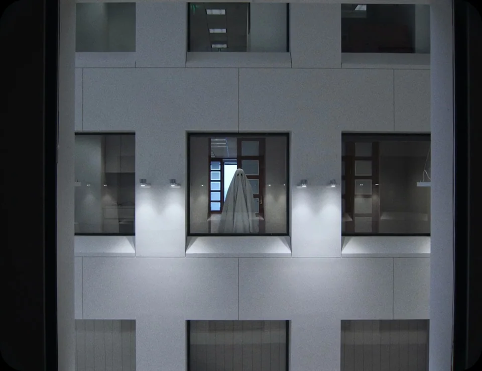 A Ghost Story (2017) movie still: wide — A modern building atrium with multiple levels of glass-paned office windows in stone walls. A single figure…