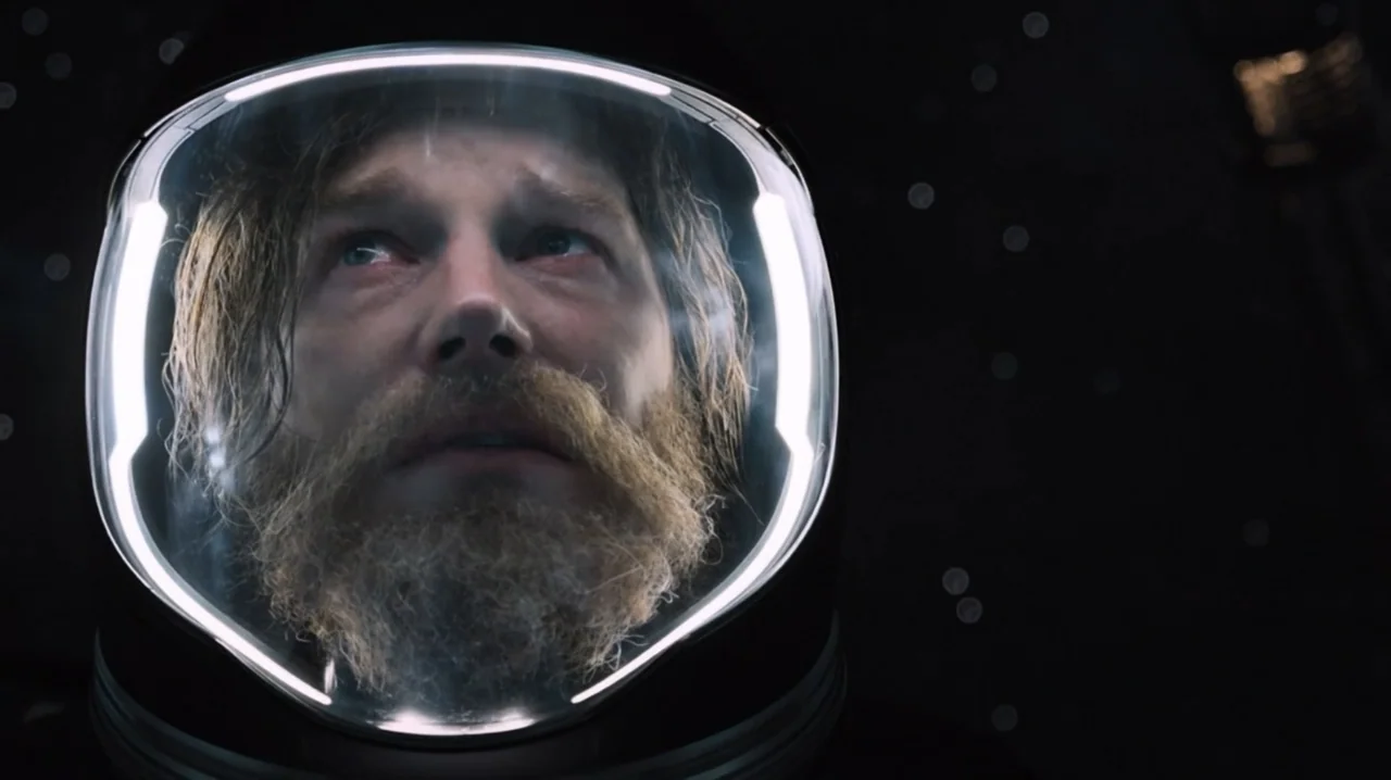 Passengers (2016) movie still: closeup — A dark void of outer space surrounds a man wearing a pressurized astronaut suit. He looks upward through…