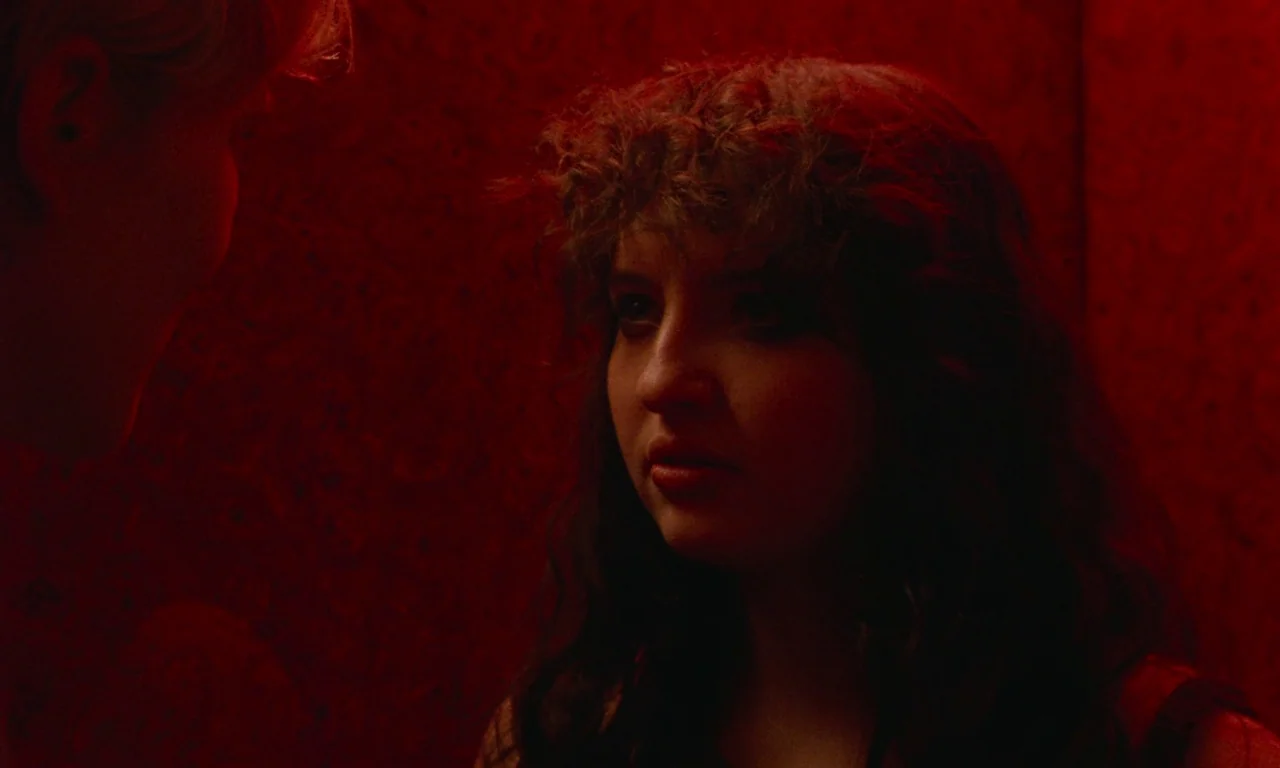 Blue Jean (2022) movie still: closeup — A confined elevator interior features walls covered in deep red textured wallpaper. A woman with curly brown…