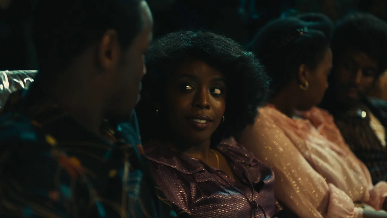 Lover’s Rock (2020) movie still: medium — An interior space occupied by people seated in dark rows. A woman turns her head to look toward a man…