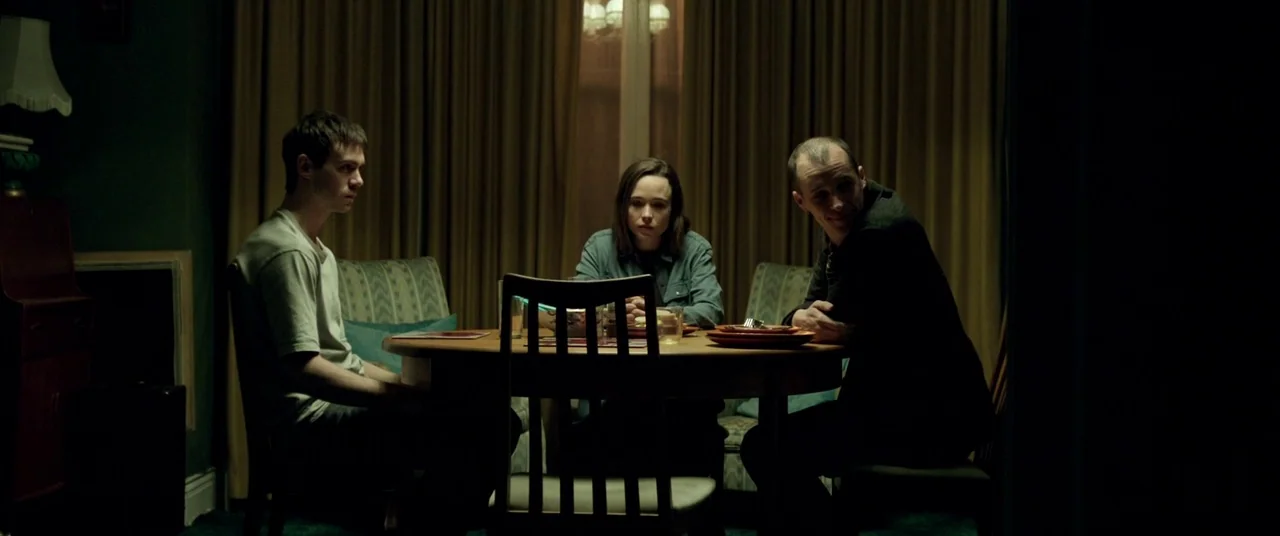 The Cured (2017) movie still: medium — A dark, dimly lit dining area features a circular wooden table draped with gold curtains. Three people sit…