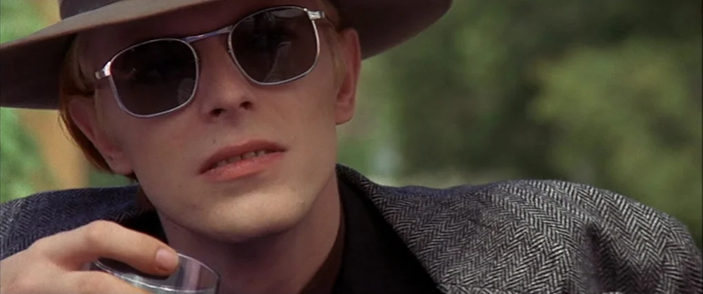 The Man Who Fell To Earth (1976) movie still: closeup — A man stands outdoors with soft foliage blurred in the background. The man holds a glass…
