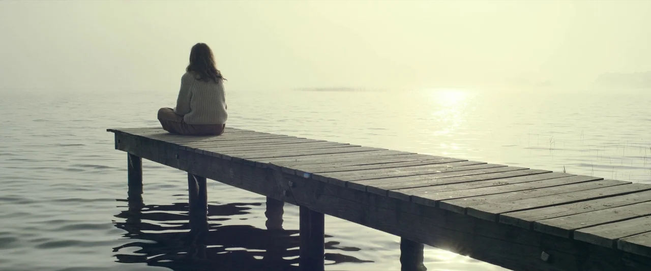 The Pod Generation (2023) movie still: wide — A wooden pier extends into a foggy lake under a bright, overcast sky. A woman sits on the end of the…