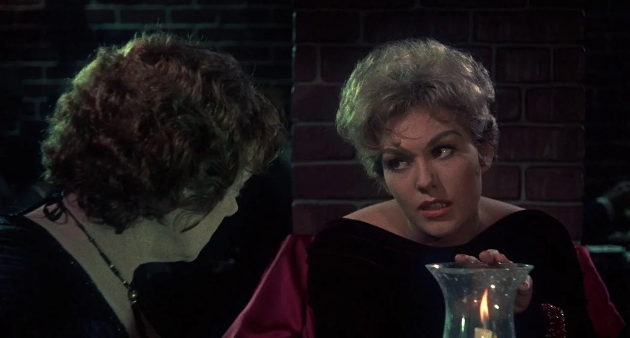 Bell Book and Candle (1958) movie still: medium — Two women are seated at a table in a dimly lit restaurant with brick walls. One woman with blonde…