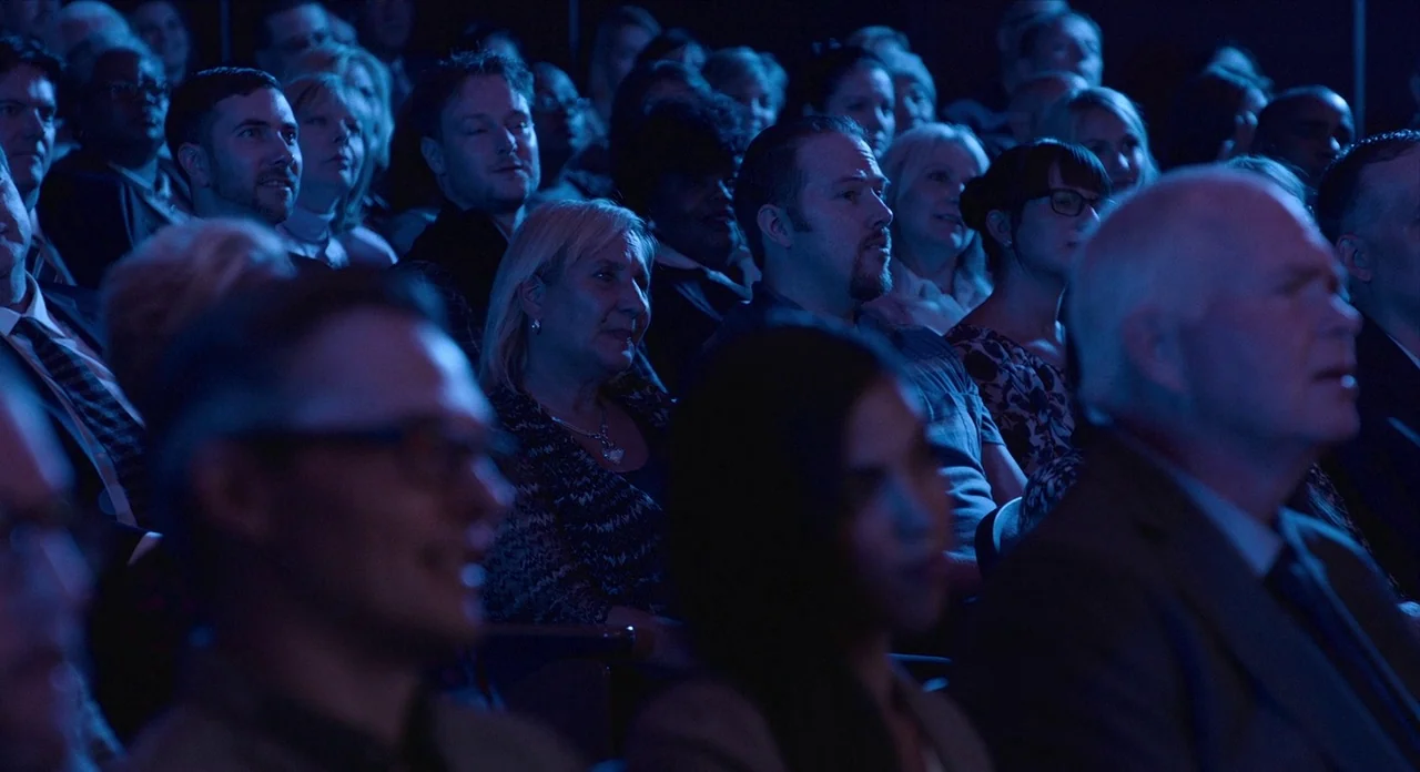 Where’d You Go Bernadette (2019) movie still: medium — A dark auditorium holds a seated crowd of people facing a light source. The audience watches…