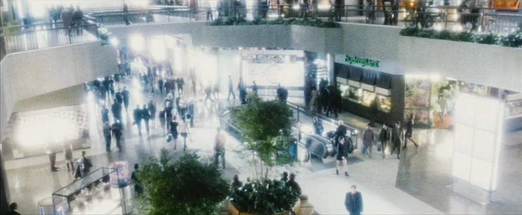 Minority Report movie still: wide — A multi-level shopping mall interior features sleek concrete architecture and indoor greenery planters. Many…
