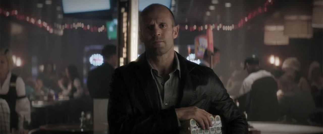 Wild Card (2015) movie still: medium — A dimly lit casino interior with patrons blurred in the background. A man stands in the foreground holding a…