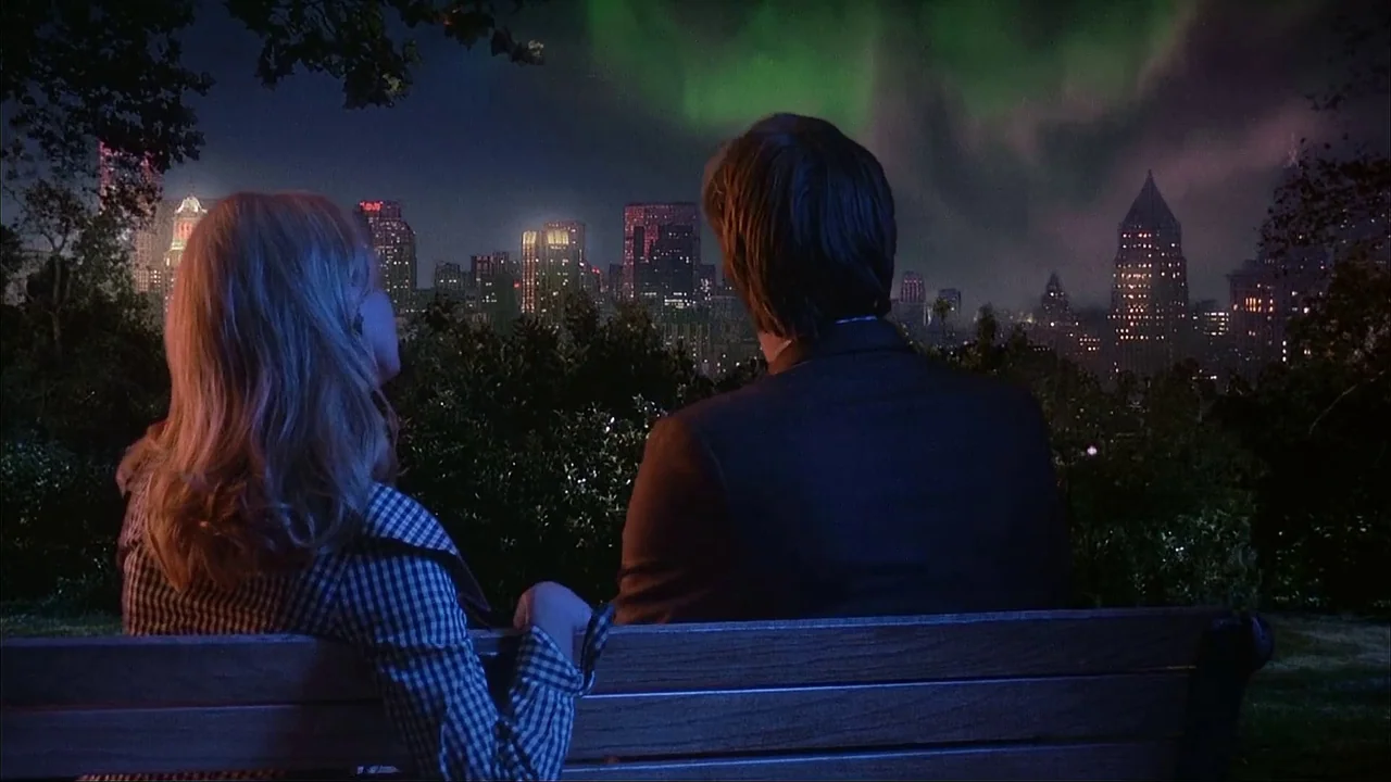 The Mask (1994) movie still: medium — A wooden park bench sits in the foreground with a sprawling illuminated city skyline behind. A man and a woman…