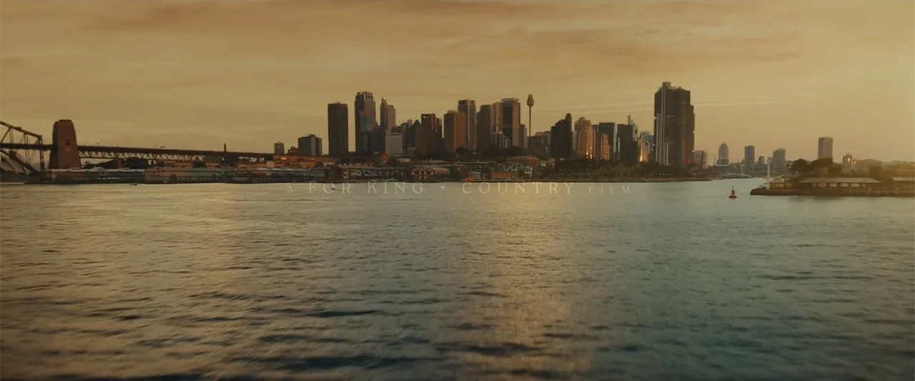 Unsung Hero (2024) movie still: establishing — A wide view of a large harbor with a sprawling city skyline visible afar. The orange light of golden…