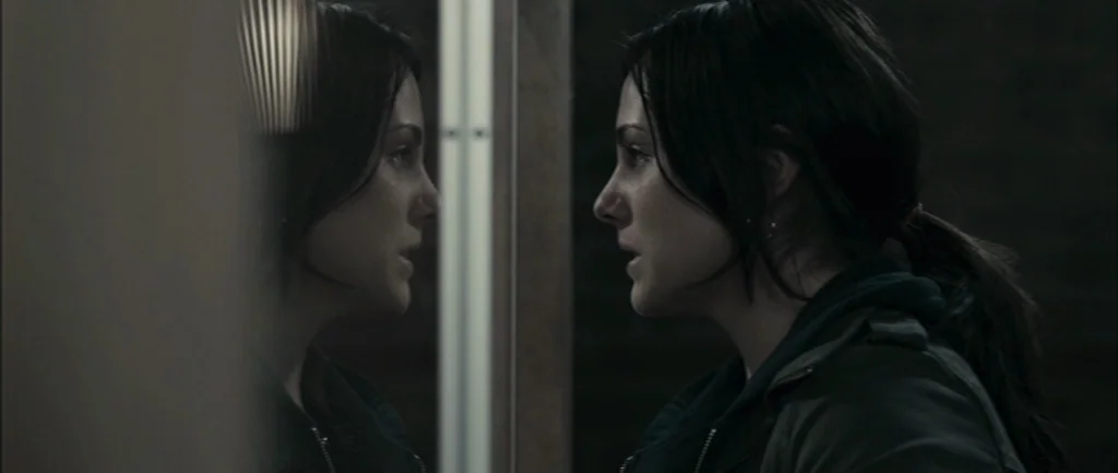 Pontypool (2008) movie still: closeup — A dimly lit space features a woman looking into a vertical wall-mounted mirror. Her profile is visible on…