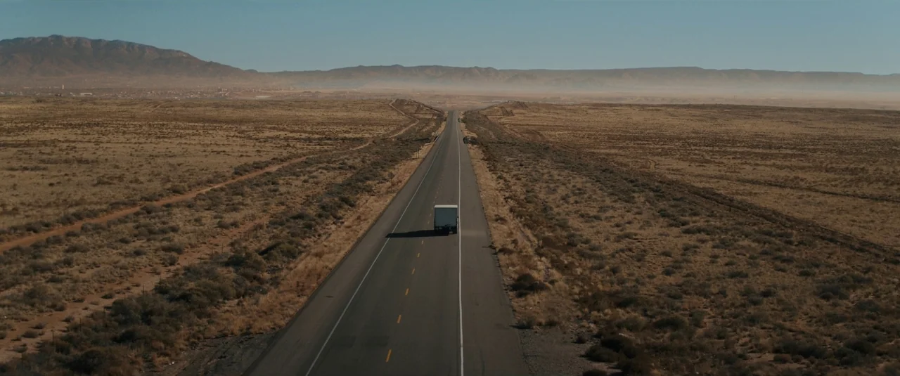 Intrusion (2021) movie still: establishing — A vast, arid desert landscape is bisected by a long, straight two-lane highway. A white truck drives…