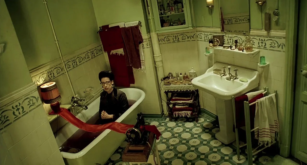 Delicatessen (1991) movie still: wide — A vintage tiled bathroom with a green and white color palette features patterned flooring. A woman sits…