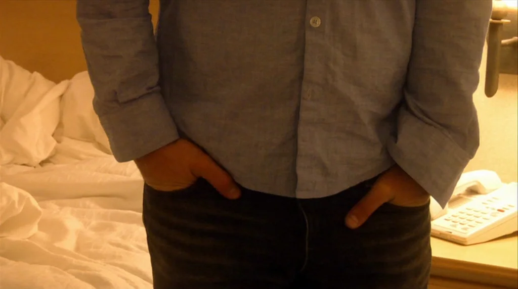 Humpday (2009) movie still: medium — A hotel room with a bed and a white landline telephone visible on a stand. A person wears a blue button-down…