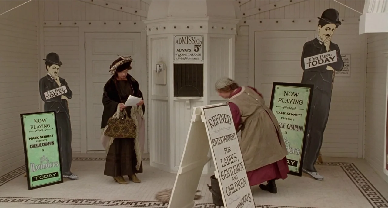 Chaplin (1992) movie still: wide — A white-walled 1920s cinema ticket booth area features classic signage and movie posters. Two women in vintage…