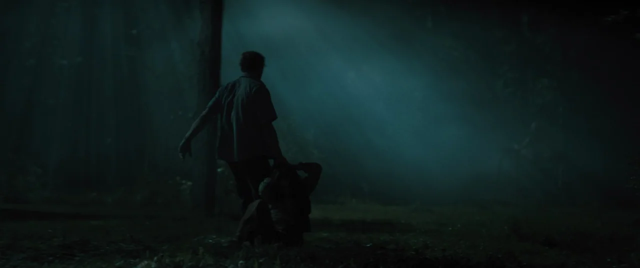 No One Will Save You (2023) movie still: wide — A dark, misty woodland clearing is illuminated by piercing beams of moonlight from above. A man…