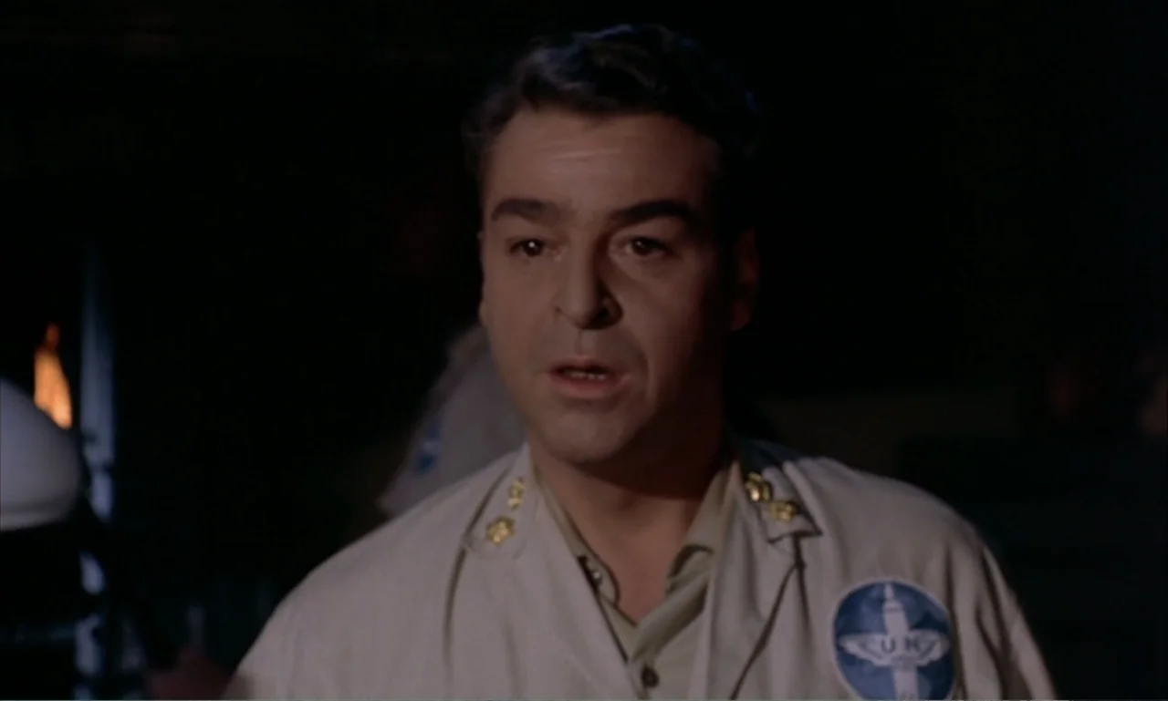 Journey to the Seventh Planet (1962) movie still: closeup — A dark indoor space features a man centered in the foreground. He wears a tan uniform…