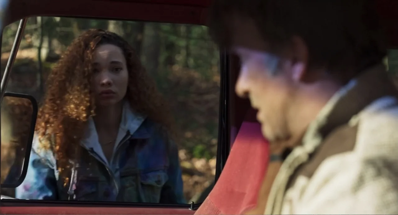 Salvation (2025) movie still: medium — A woman stands outside the driver-side window of a car in a wooded area. The driver sits inside the vehicle…