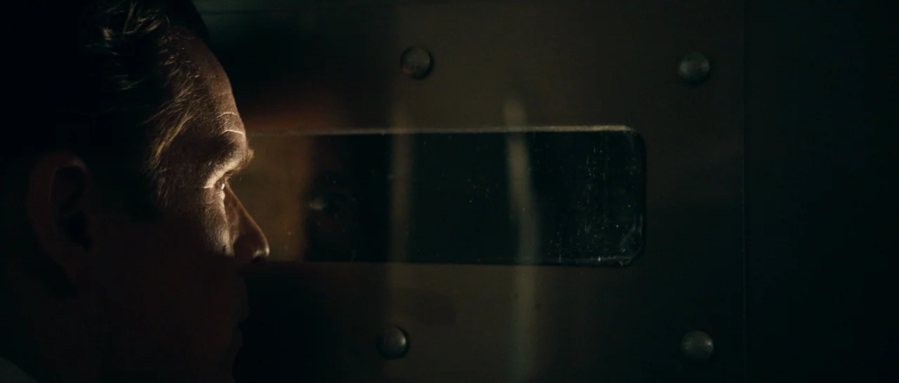 The Purge (2013) movie still: closeup — A dark, heavy metal door features a small observation window with rounded corners. A man's face is…