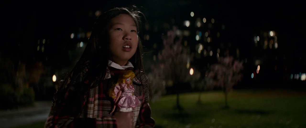 Colossal (2016) movie still: medium — A nighttime park setting with blurred distant city lights in the dark background. A young girl in a plaid coat…