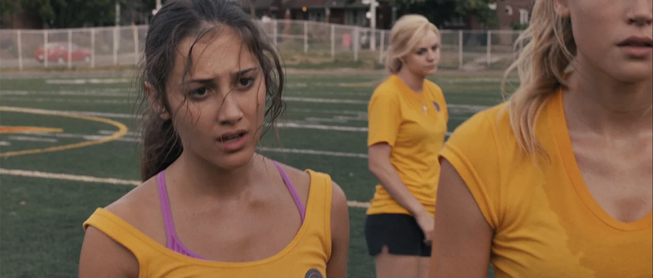 Carrie (2013) movie still: medium — A green artificial turf athletic field features white boundary lines and a perimeter chain-link fence. Three…
