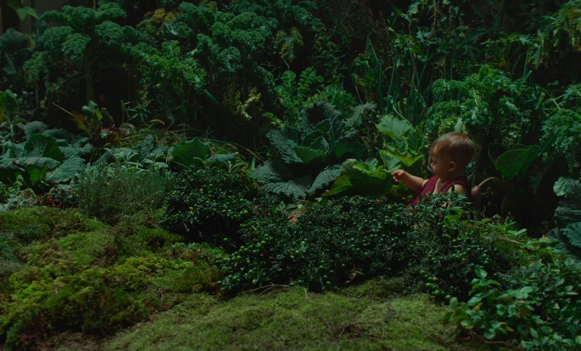 High Life (2018) movie still: wide — A dense vegetable patch filled with kale, herbs, and low berry bushes. A small child sits amidst the greenery,…