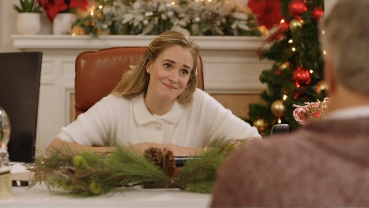 Loving Christmas (2022) movie still: medium — A festive office setting features a decorated fireplace mantle and a Christmas tree behind a desk. A…