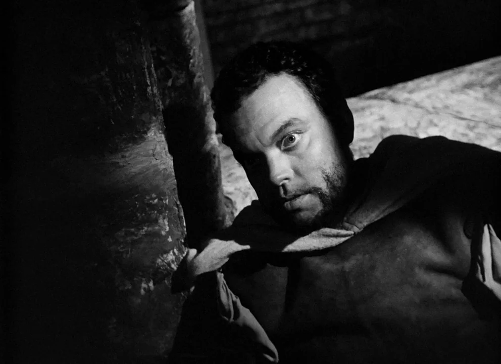 Othello (1952) movie still: closeup — A dark, textured interior setting with high-contrast lighting cast against a rough masonry pillar. A man gazes…