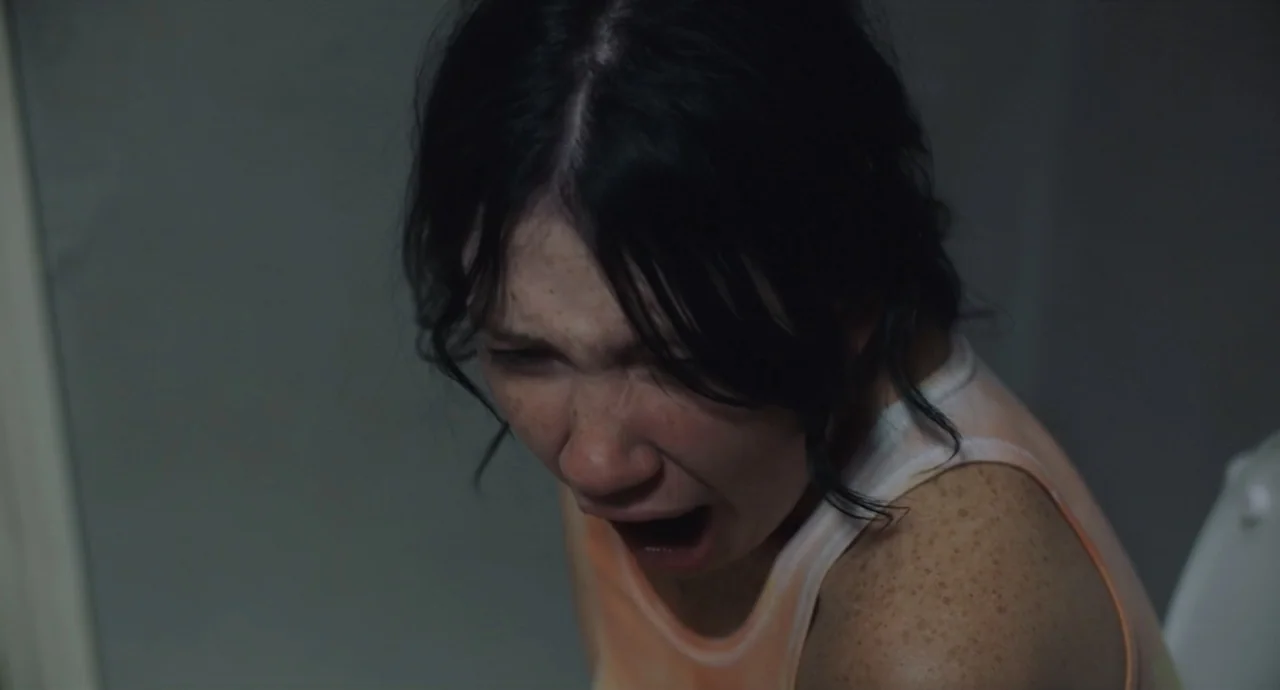 Pleasure (2021) movie still: closeup — A dimly lit indoor space with grey walls and a white surface visible. A young woman with freckled skin cries…
