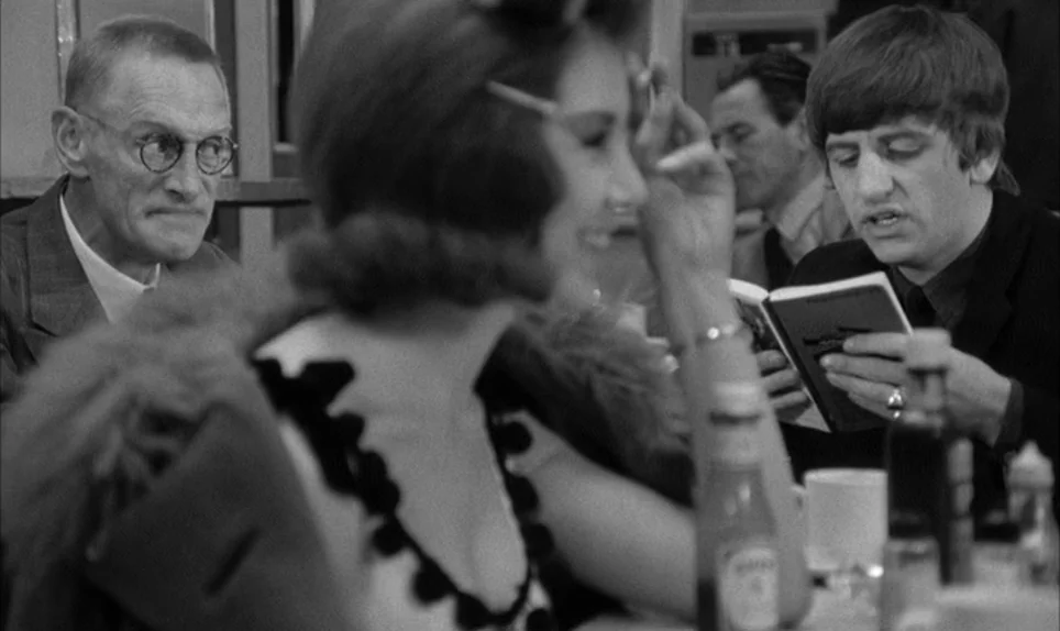 A Hard Days Night (1964) movie still: medium — A diner booth occupied by several patrons sitting at a table with condiments. A young man reads a…