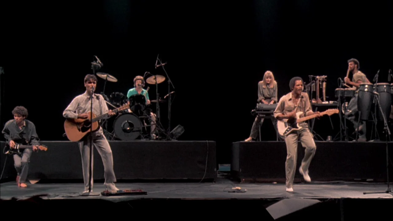 Stop Making Sense (1984) movie still: wide — A musical group performs on a minimalist stage against a solid black backdrop. Members play electric…
